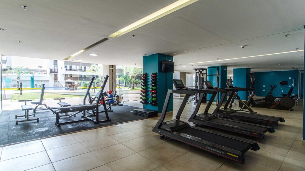 Academia e Fitness Center – Venice Park Academia moderna do Venice Park equipada com esteiras da marca Movement, área de musculação com bancos de supino, halteres organizados em suportes verticais nas colunas azuis e vista para a área externa e piscina.