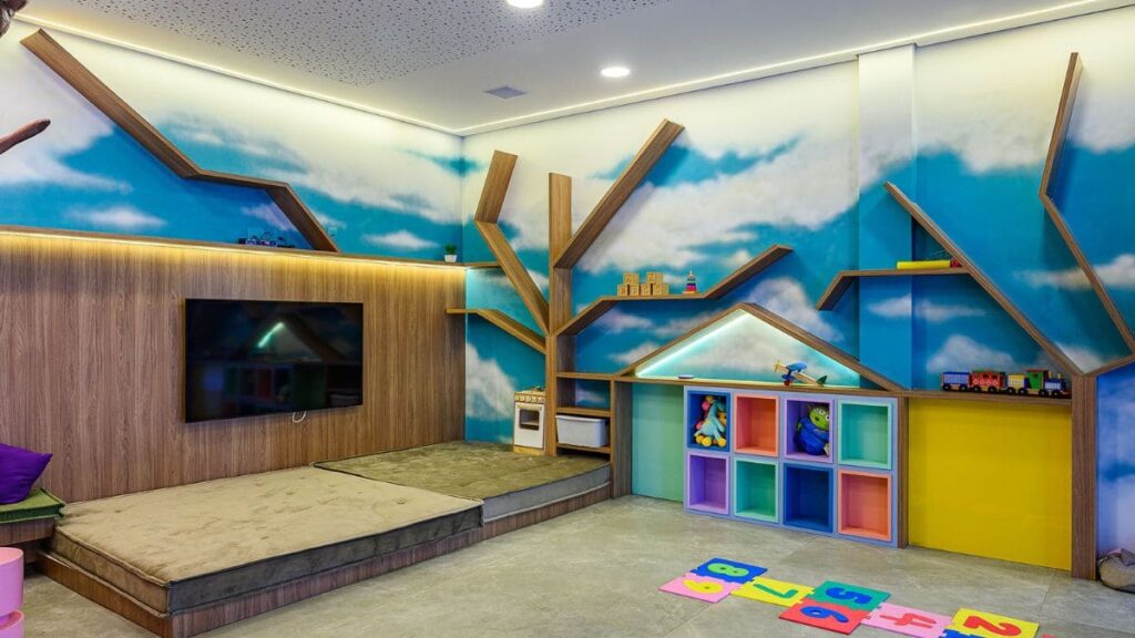 Brinquedoteca lúdica e decorada com painel de TV em madeira, prateleiras em formato de árvore, colchões no chão e paredes com estampa de céu em um Apartamento 4 Quartos Noroeste - Residencial Singular.