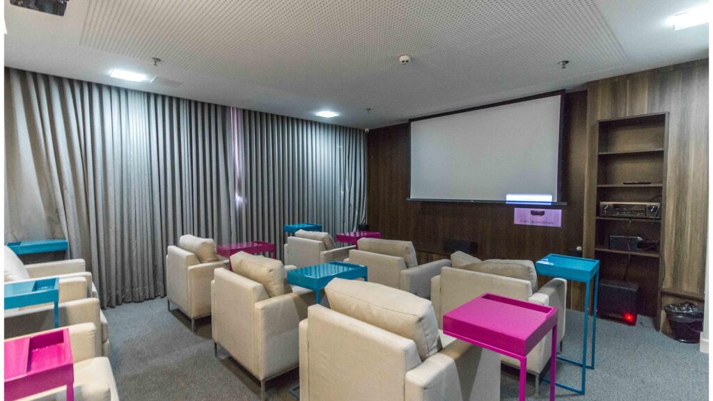 Cine Lounge e Entretenimento – Venice Park Espaço de cinema no Venice Park com poltronas individuais estofadas em tom bege, mesinhas de apoio coloridas em azul e rosa, carpete cinza, cortinas black-out e projetor instalado no teto.