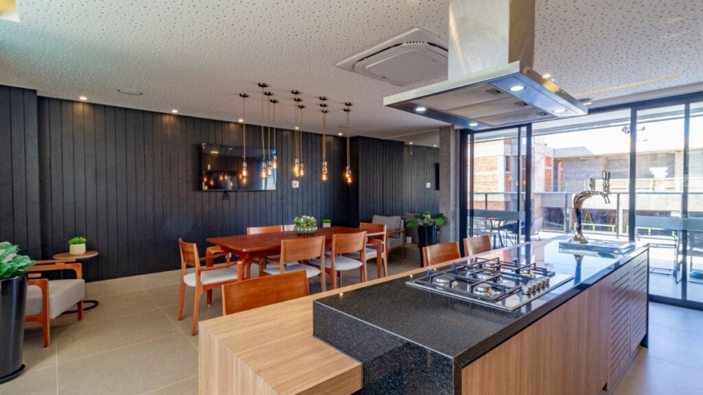 Área gourmet moderna de um condomínio com ilha central, cooktop, coifa de inox e mesa de jantar em madeira, ideal para quem busca um apartamento 2 quartos Noroeste.