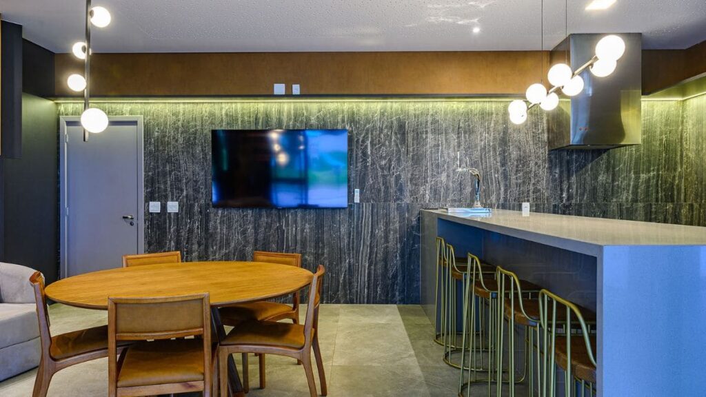 Espaço Gourmet luxo com mesa de jantar oval em madeira, bancada gourmet com banquetas de metal dourado e painel de TV em pedra em um Apartamento 4 Quartos Noroeste - Residencial Singular.