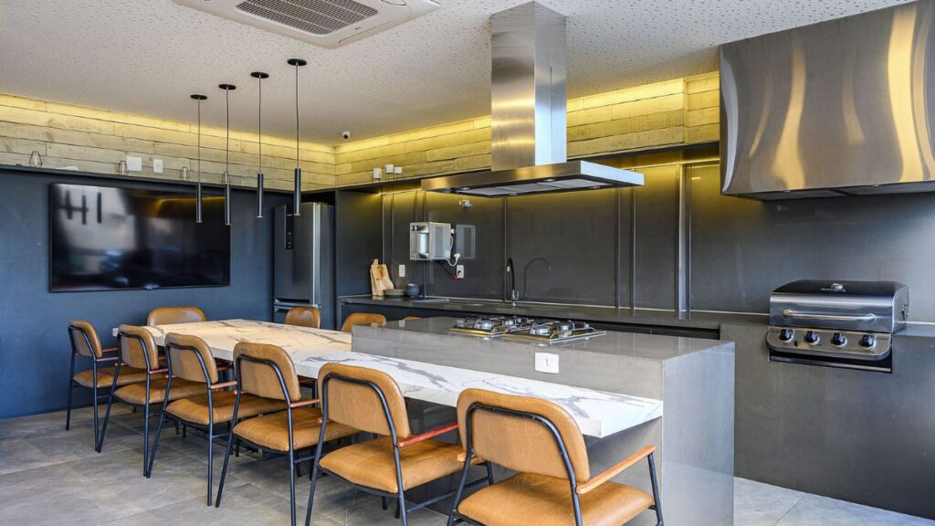 Espaço gourmet de luxo com ilha central em mármore, churrasqueira embutida, coifa de inox e mesa de jantar integrada em um Apartamento 4 Quartos Noroeste - Residencial Singular.