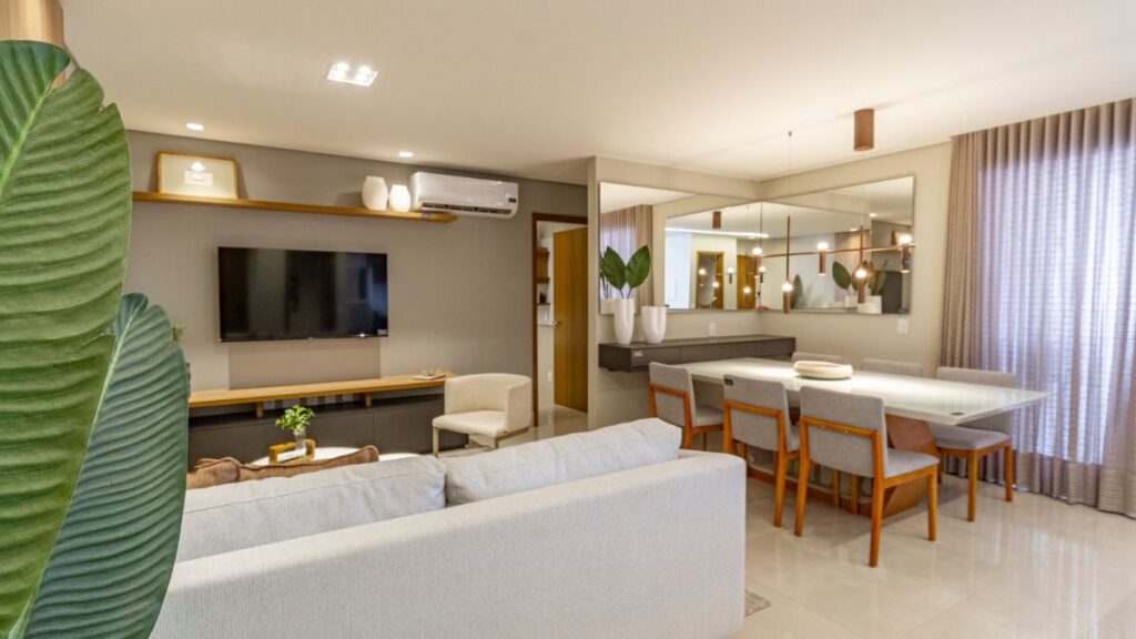 Sala de estar e jantar integrada do Residencial Unité em Águas Claras, apresentando sofá cinza, painel de TV moderno, mesa de jantar para seis pessoas com espelhos ao fundo e iluminação elegante.