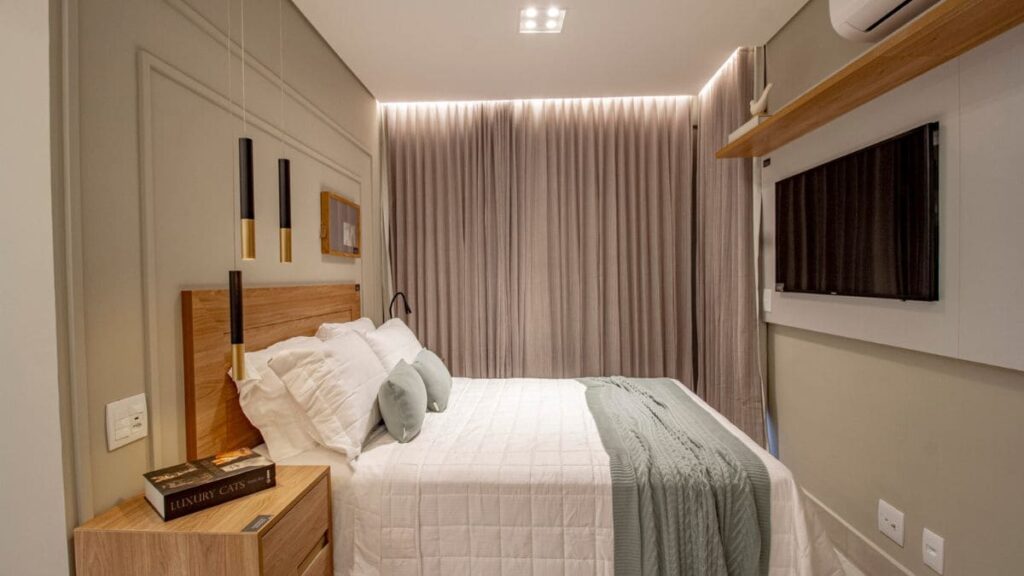 Suíte principal decorada do Residencial Unité em Águas Claras, apresentando cama de casal com enxoval branco e verde, painel de madeira com luminárias pendentes modernas e TV instalada à frente.