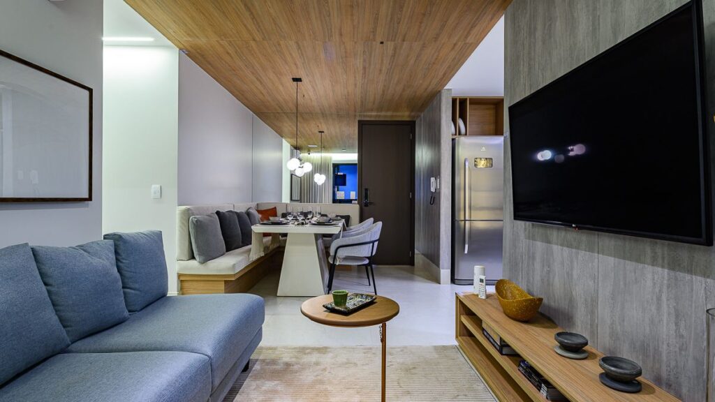 Sala de estar integrada com sofá azul, painel de TV amadeirado e vista para a mesa de jantar em um apartamento 2 quartos Noroeste.