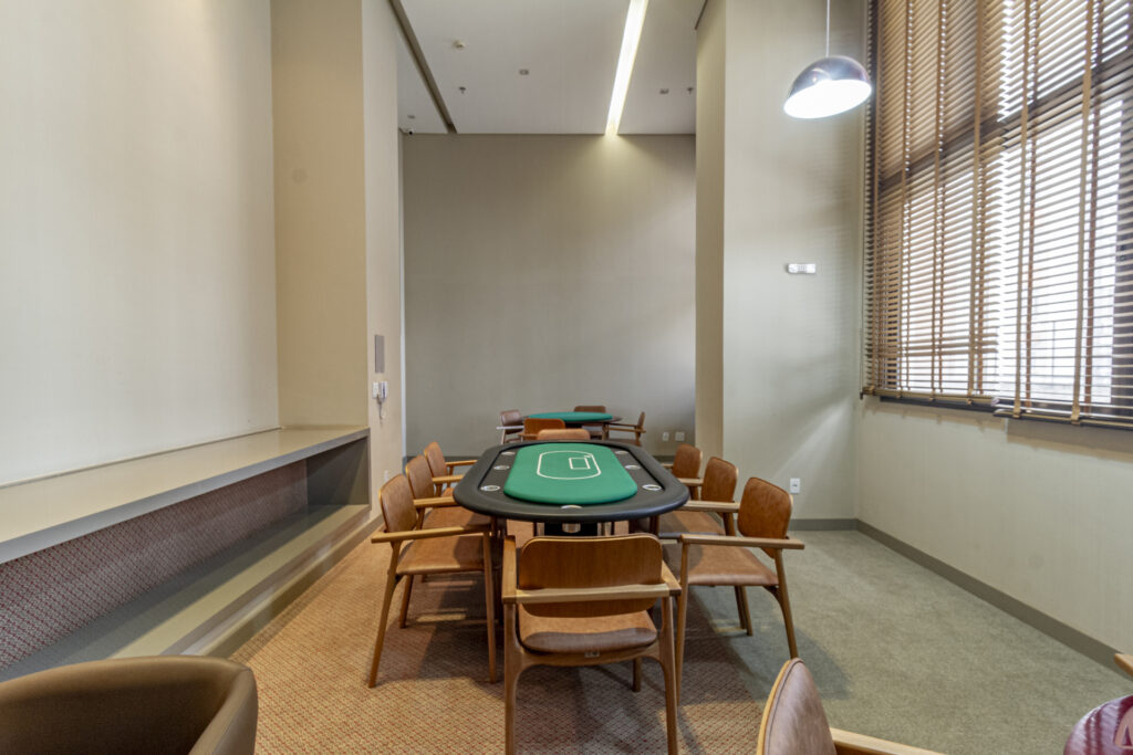 Sala de Poker e Jogos de Mesa – Venice Park Sala de jogos do Venice Park com mesa profissional de poker para oito pessoas, tampo verde, cadeiras de madeira com estofado marrom, iluminação pendente e janelas amplas com persianas horizontais.