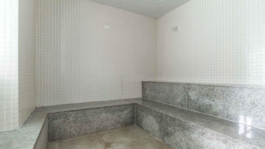 Sauna a Vapor – Venice Park Interior de uma sauna a vapor no Venice Park com revestimento de pastilhas brancas nas paredes e bancos em dois níveis feitos de granito cinza.
