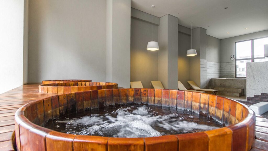 SPA com Ofurôs e Área de Descanso – Venice Park Área de SPA e bem-estar do Venice Park com dois ofurôs circulares de madeira e água borbulhando no primeiro plano, dispostos sobre um deck de madeira em um ambiente interno com cadeiras reclináveis brancas ao fundo, paredes cinzas e luminárias pendentes modernas.