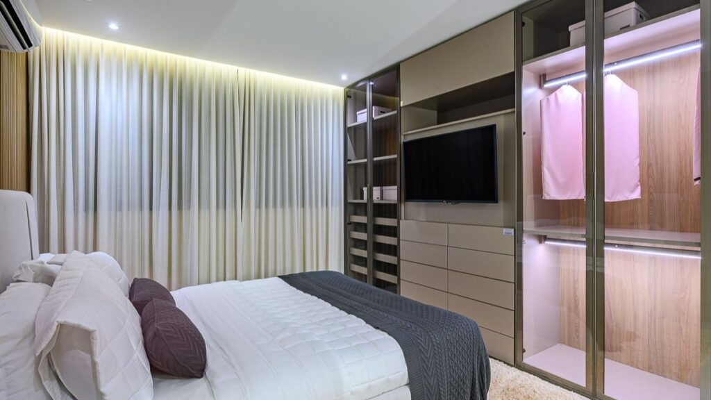 Quarto de casal decorado em um apartamento 2 quartos Noroeste, com armários planejados com portas de vidro, iluminação embutida e cama com enxoval branco.