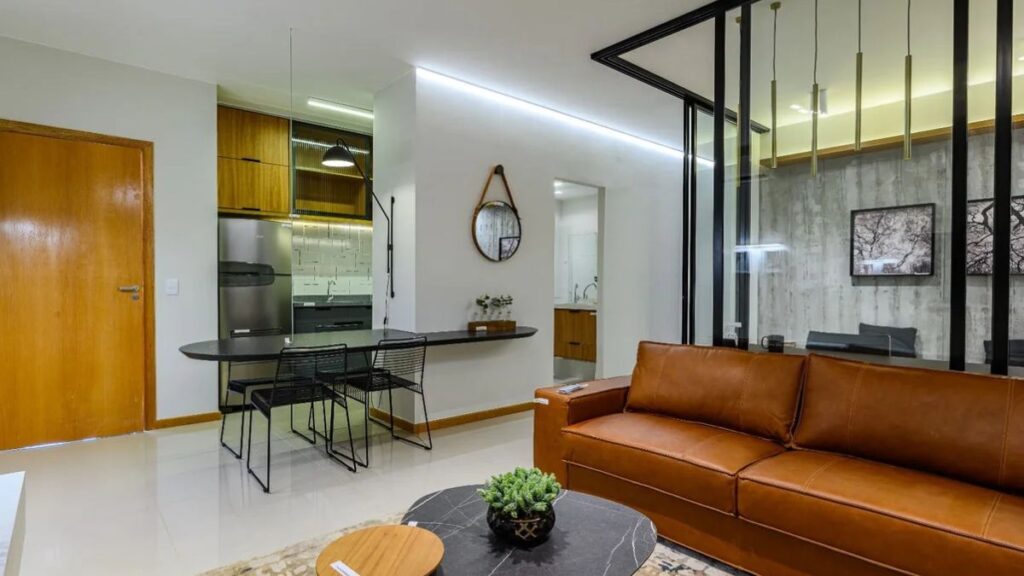 Fotografia de uma sala de estar decorada no Portal do Parque II. O ambiente integrado apresenta um sofá de couro caramelo, mesa de centro de mármore escuro e um tapete estampado. Ao fundo, uma cozinha americana com armários de madeira e bancada preta com banquetas. Uma divisória de vidro com moldura preta separa a sala de um escritório moderno.