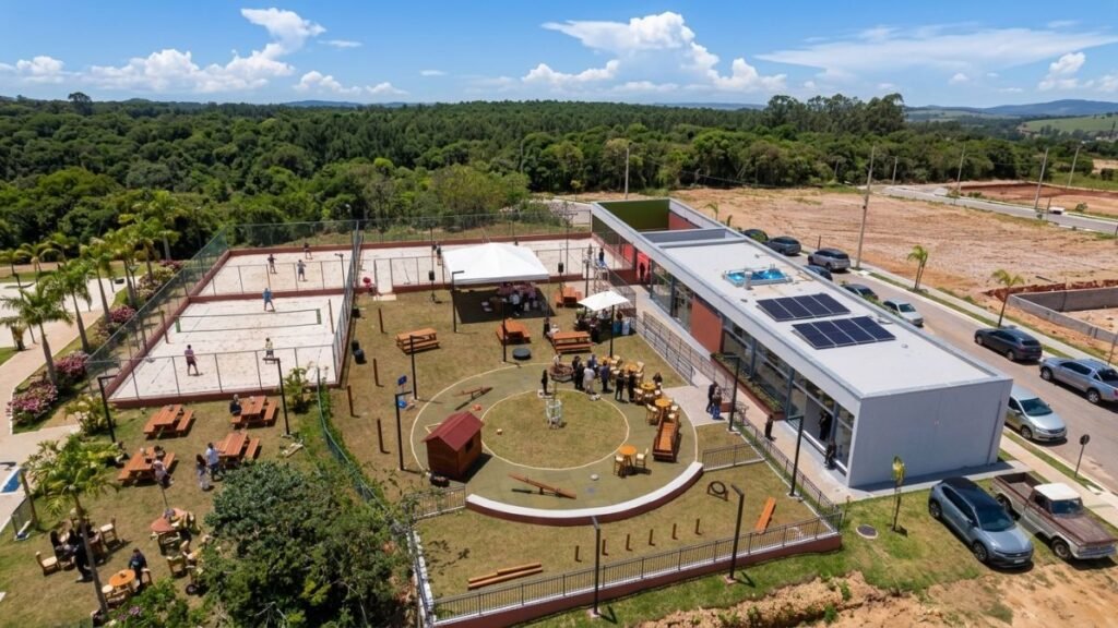Vista aérea de uma moderna área de lazer e clube de condomínio fechado Le Grand Jardin, exibindo duas quadras de areia para futevôlei ou beach tennis, parquinho infantil circular com gangorras, quiosques com mesas, e um clube contemporâneo com painéis solares no teto, rodeado por uma imensa área verde preservada, ideal para quem busca um Lote Jardim Botânico.