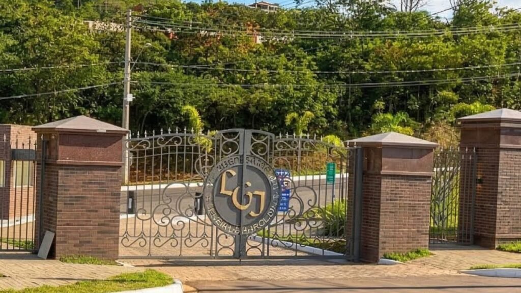 Vista frontal da imponente portaria de acesso ao condomínio fechado Le Grand Jardin, exibindo portões de ferro trabalhado com brasão central e pilares rústicos de tijolos, cercada por uma densa vegetação natural que caracteriza a busca por um Lote Jardim Botânico.