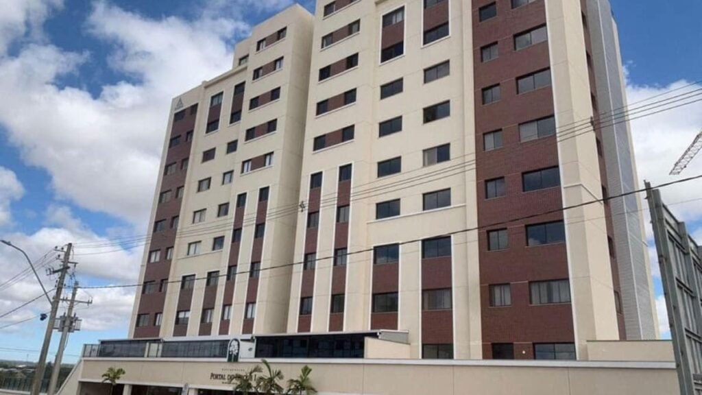 Fotografia em ângulo contrapicado mostrando a fachada de um edifício residencial de múltiplos andares com revestimento em pastilhas marrons e pintura bege. Na base térrea, identifica-se a identificação visual e a entrada do condomínio Portal do Parque I.