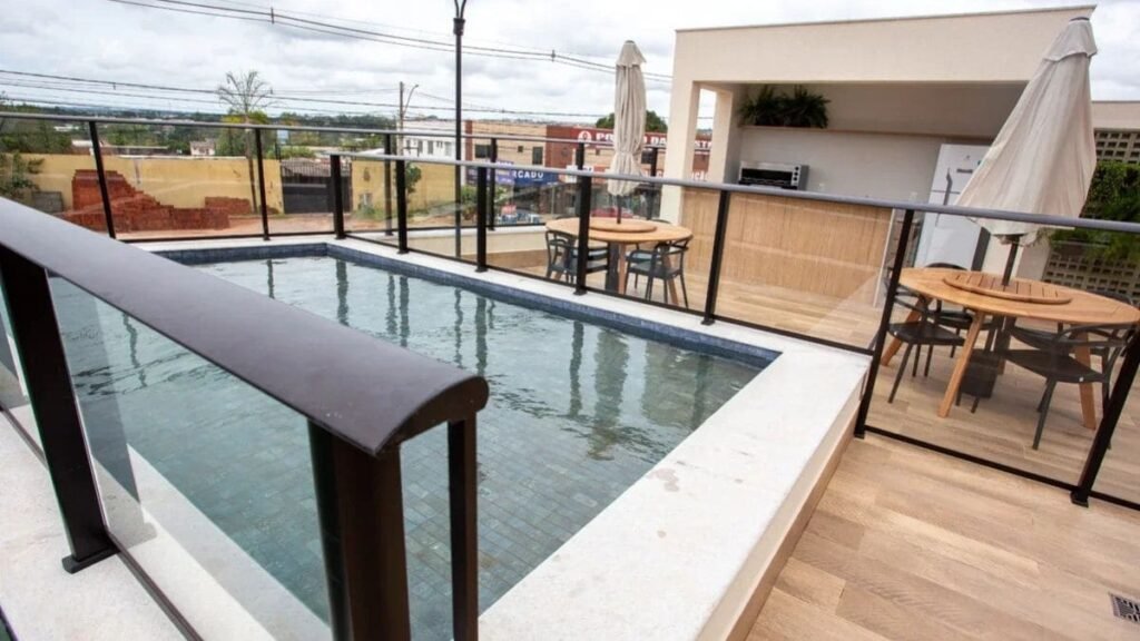 Fotografia em perspectiva de uma área de lazer elevada em uma cobertura de condomínio, destacando uma piscina de pastilhas escuras cercada por guarda-corpo de vidro. Ao lado, há um deque com mesas de madeira e ombrelones, ilustrando a infraestrutura do Portal do Parque I.
