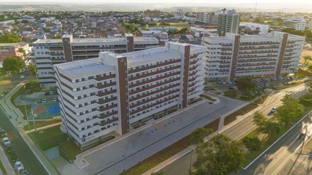 Fotografia aérea panorâmica exibindo três grandes blocos de edifícios residenciais alinhados, com fachadas em tons de branco e marrom. A imagem mostra as vias de acesso, áreas pavimentadas e o entorno urbano do Residencial Quadra Parque Gama sob a luz do dia.